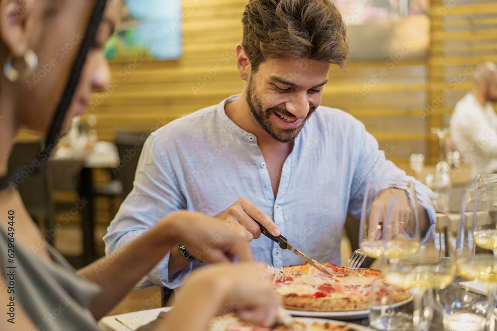 Man Enjoying Pizza in Courtyard Restaurant - Young man smiles cutting his pizza, blurred close-up of an African American woman in the foreground.