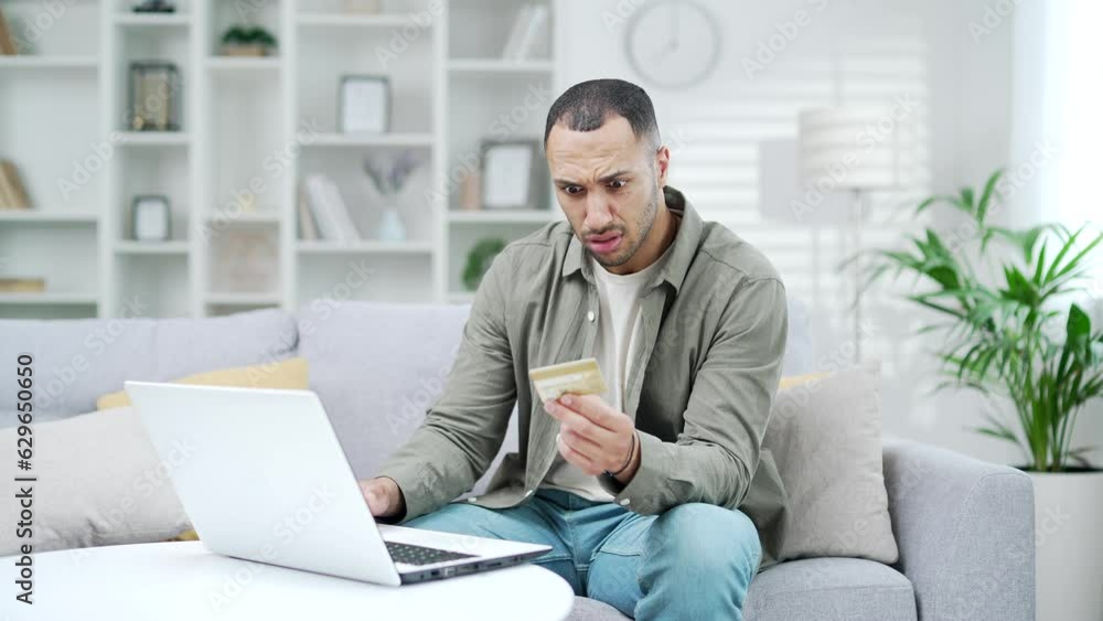 Sad male discovered fraud by entering credit card number on laptop while sitting at couch in home office. A disappointed young adult mixed race male saw that money had been stolen from their account Sad male discovered fraud by entering credit card number on laptop while sitting at couch in home office. A disappointed young adult mixed race male saw that money had been stolen from their account