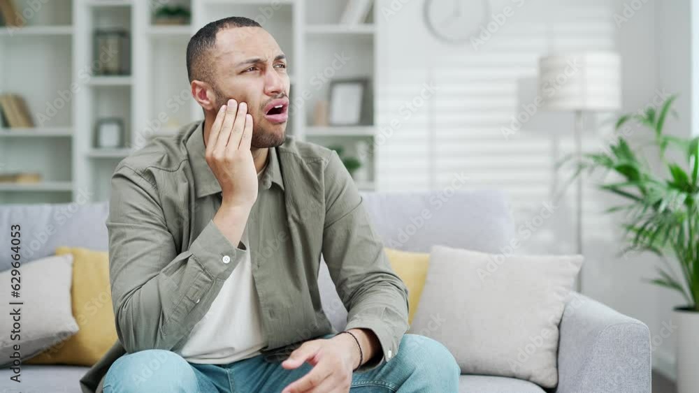 Sad handsome male suffering from toothache while sitting at home in living room. Young adult guy suffer from terrible painful tooth ache, sensitive teeth cavities Indoor in a modern interior Sad handsome male suffering from toothache while sitting at home in living room. Young adult guy suffer from terrible painful tooth ache, sensitive teeth cavities Indoor in a modern interior