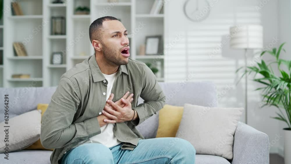 Mixed race man with heart pain holding his chest. Heart Attack Adult male has severe chest pain aches. Myocardial Infarction. Pressure while sitting on a couch at home in the living room alone indoor Mixed race man with heart pain holding his chest. Heart Attack Adult male has severe chest pain aches. Myocardial Infarction. Pressure while sitting on a couch at home in the living room alone indoor
