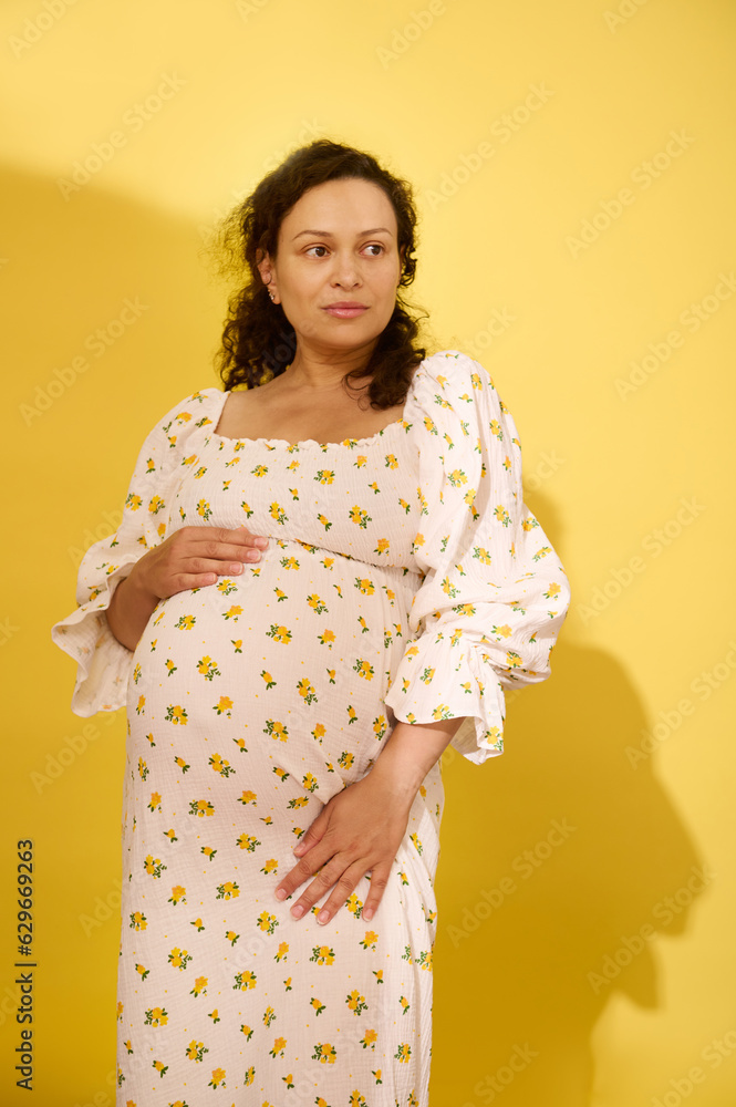 Beautiful curly haired pregnant woman in summer sundress, holding her belly, looking aside, isolated yellow background Beautiful curly haired pregnant woman in summer sundress, holding her belly, looking aside, isolated yellow background