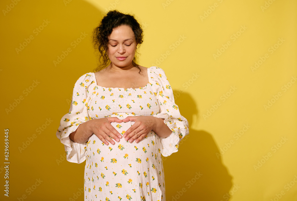 Multi-ethnic curly haired pregnant woman making a heart shape on her big belly, expressing love, care and tenderness Multi-ethnic curly haired pregnant woman making a heart shape on her big belly, expressing love, care and tenderness
