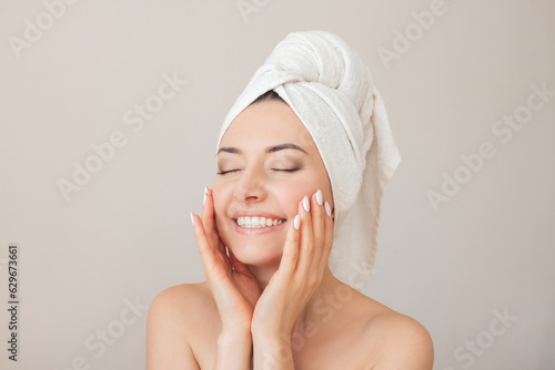 portrait of a young happy beautiful naked woman with closed eyes with a towel on her head smiling and gently touching her face with her hands. Isolated on a beige background
