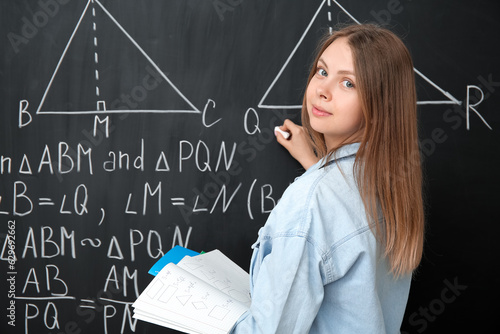 Young math teacher with copybooks writing on blackboard in classroom