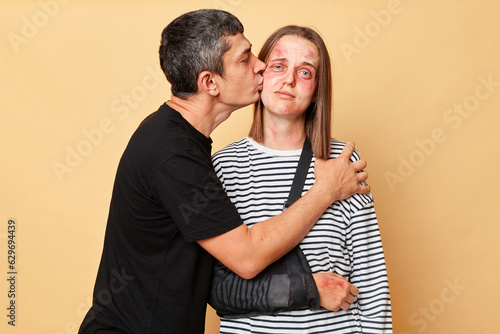 Man and woman with bruise isolated over beige background, tyrant husband asks for forgiveness from wife after brutal beating kissing her cheek. photo