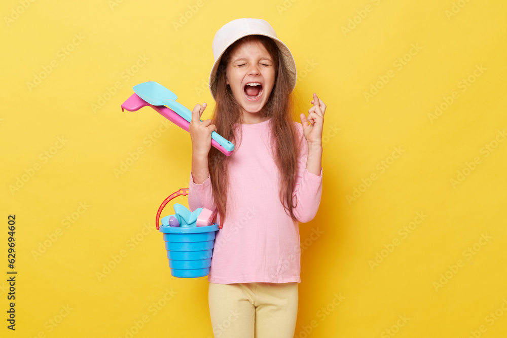 Cheerful joyful happy little girl wearing casual clothing and panama holding sandbox toys and bucket isolated over yellow background crossing fingers making wish. Cheerful joyful happy little girl wearing casual clothing and panama holding sandbox toys and bucket isolated over yellow background crossing fingers making wish.