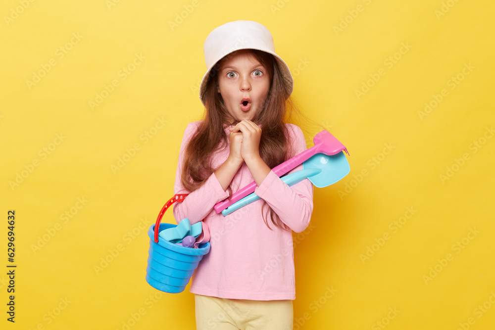 Surprised little girl wearing casual clothing and panama holding sandbox toys and bucket isolated over yellow background posing with rake and scoop keeps mouth open. Surprised little girl wearing casual clothing and panama holding sandbox toys and bucket isolated over yellow background posing with rake and scoop keeps mouth open.