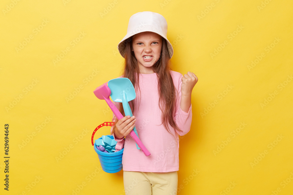 Angry little girl wearing casual clothing and panama holding sandbox toys and bucket isolated over yellow background standing with clenched teeth someone took her scoop and rake at playground Angry little girl wearing casual clothing and panama holding sandbox toys and bucket isolated over yellow background standing with clenched teeth someone took her scoop and rake at playground