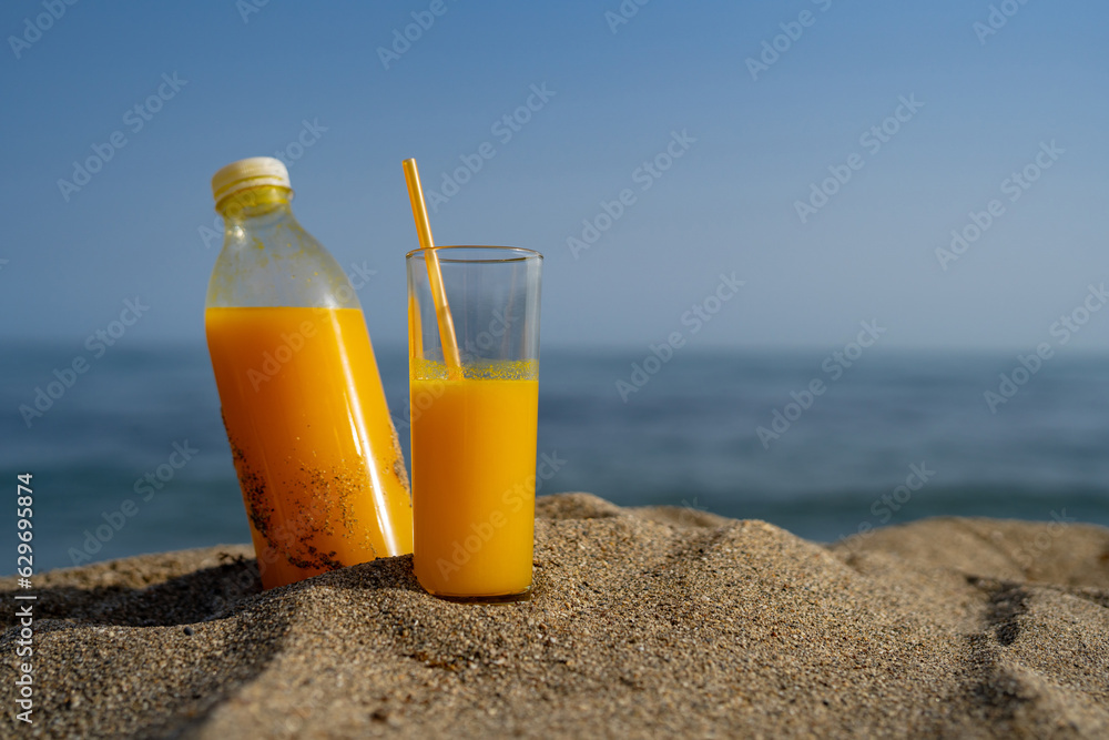a bottle and a glass of orange juice stand on the sand against the background of the sea