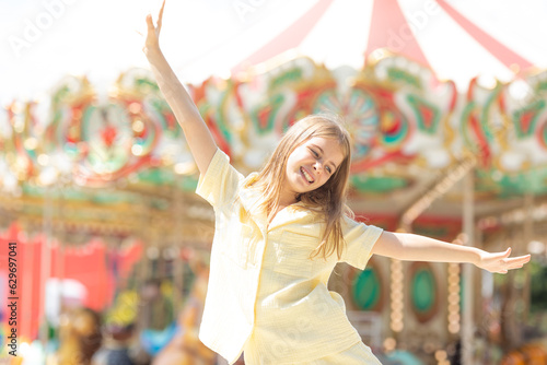 Freedom. Adorable student blonde girl in summer yellow clothes at amusement park with arms raised high . Cute girl stands on the background of the carousel and laughs. Entertainment concept