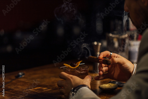 Man smoking a cigar and having an old-fashioned cocktail served on a wooden bar table