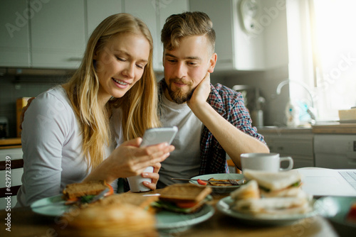 Canvastavla Young couple using a smart phone while having breakfast in the kitchen in the mo