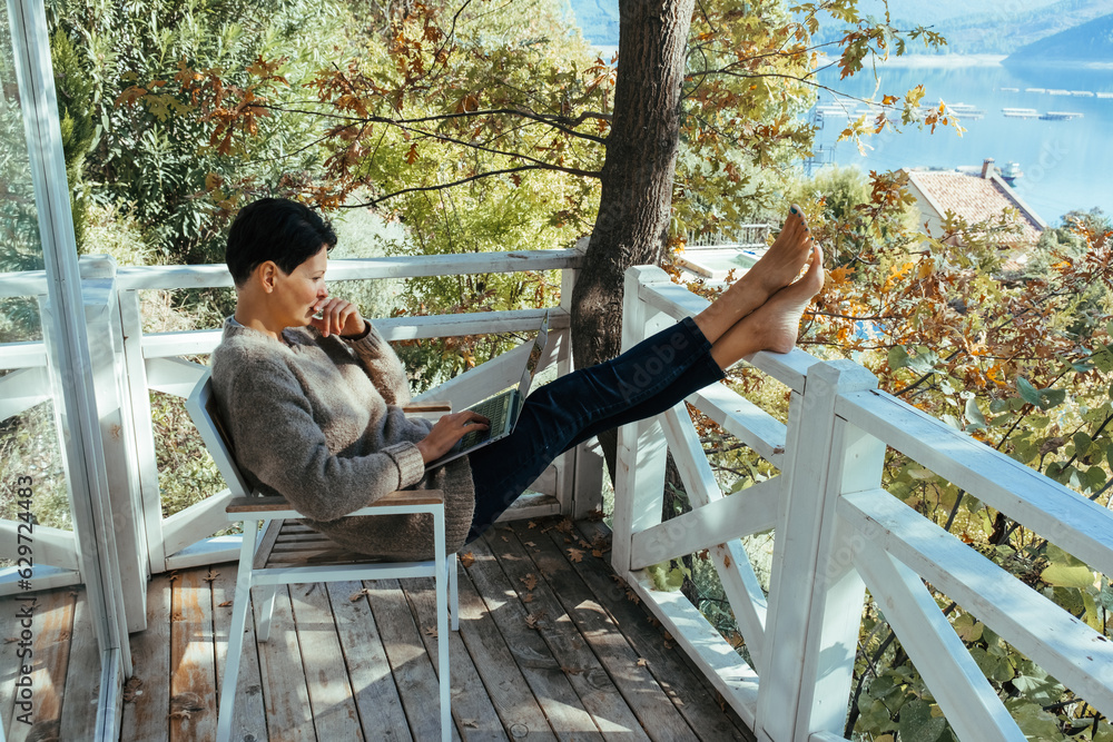 Fototapeta premium Middle-aged woman working on laptop computer sitting at table on wooden balcony of country house. High quality photo