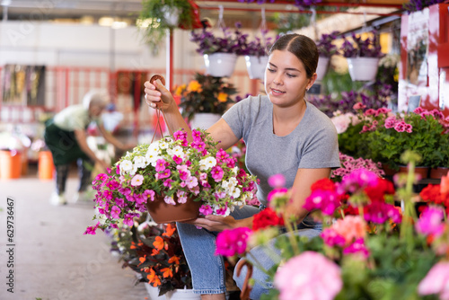 Cheerful young girl choosing ornamental plants to decorate courtyard at flower market, looking with interest at flowering petunias in hanging cache-pots