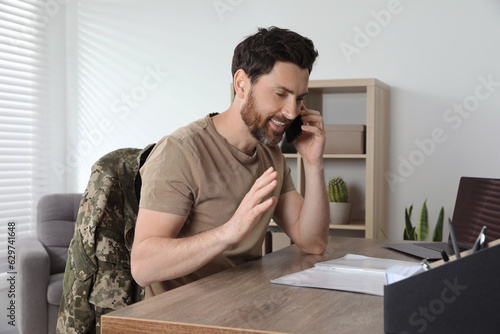 Happy soldier talking on phone at wooden table indoors. Military service