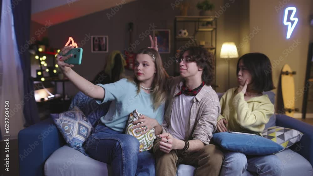 Three friends make funny selfies. The girl holds phone in her hand, grimacing, making funny faces. Virtual life in social media. Relaxation recreation at free time. Leisure with smartphone in internet Three friends make funny selfies. The girl holds phone in her hand, grimacing, making funny faces. Virtual life in social media. Relaxation recreation at free time. Leisure with smartphone in internet