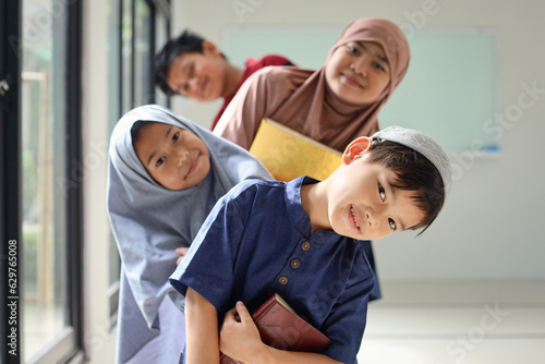 Canvas Print Happy smiling Muslim little kids holding holy book of Quran and standing in a row one after another near window