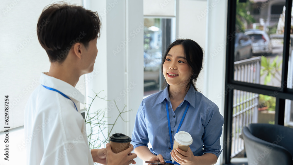 two young asian businesswomen standing chatting talking and holding coffee by the window in office