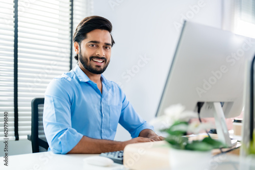 Indian professional business man, businessman working on computer, looks directly at camera and smiles friendly