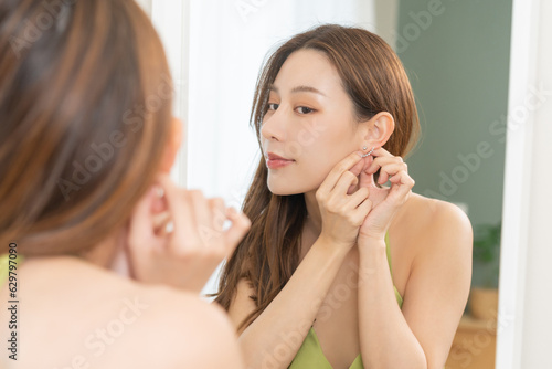 Get dress, elegant asian young woman with makeup glamour, getting ready wearing green cloth, gorgeous female putting earring or jewelry looking into the mirror at home, prepare going event night party