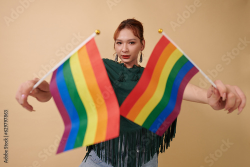 Transgender woman standing against yellow background with rainbow flags, drinking wine and looking at camera