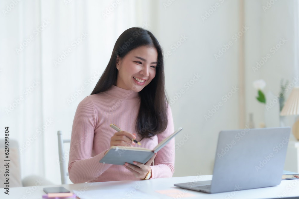 Smiling happy asian businesswoman sitting with laptop computer taking notes in office.