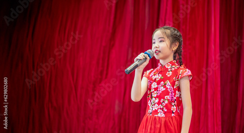 The Asian kid girl sing a song on stage at her school activity day, dress in Qipao style, red curtain background