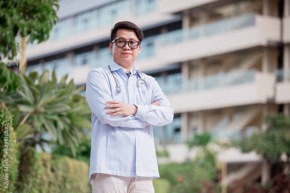 Horizontal waist-up portrait of a handsome male Arab Malay doctor, wearing white coat, standing with arms crossed outdoors on the background of clinic building. Close up, copy space Horizontal waist-up portrait of a handsome male Arab Malay doctor, wearing white coat, standing with arms crossed outdoors on the background of clinic building. Close up, copy space