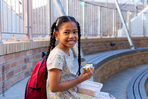 Portrait of happy biracial schoolgirl eating healthy lunch eating sandwich at elementary school