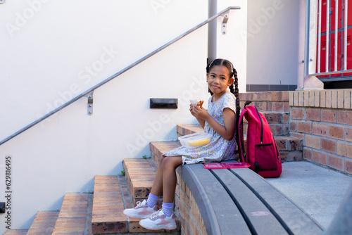 Portrait of happy biracial schoolgirl having healthy lunch eating sandwich at elementary school