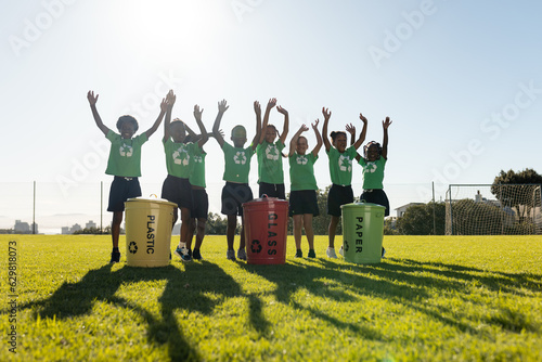 Portrait of happy diverse children collecting recycling waving in sunny elementary school field