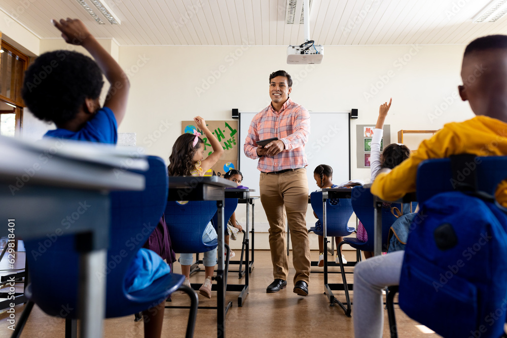 Obraz premium Diverse male teacher with tablet and elementary schoolchildren raising hands in class