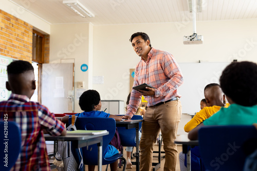Diverse male teacher with tablet and elementary schoolchildren sitting at desks in class