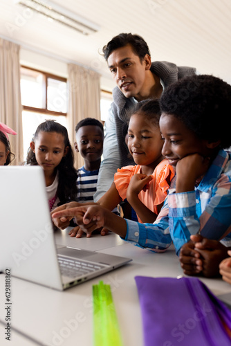 Diverse male teacher using laptop teaching children in class at elementary school