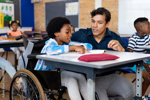 Biracial male teacher helping schoolboy sitting in wheelchair in classroom at elementary school