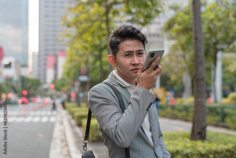 A young man talks to someone via speakerphone or using voice recognition app technology to get directions to an address on his cellphone while walking. Outdoor city scene. A young man talks to someone via speakerphone or using voice recognition app technology to get directions to an address on his cellphone while walking. Outdoor city scene.