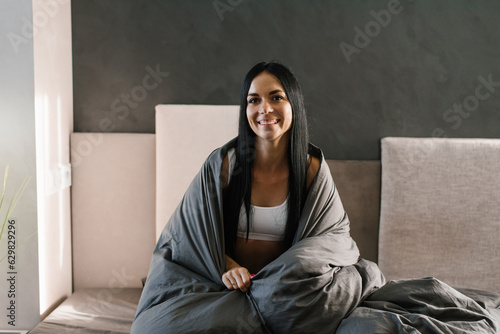 Young beautiful brunette in pajamas wrapped in a blanket sits on the bed in the morning after sleeping and smiles