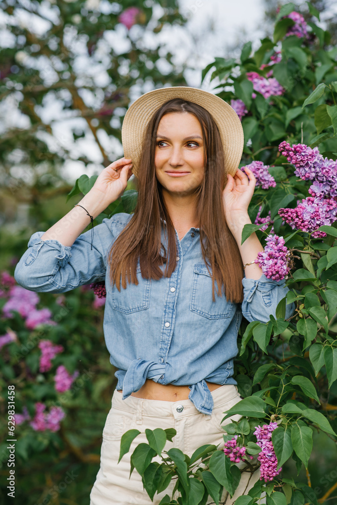 Pretty young woman in a denim shirt and a straw hat walks among the blooming lilacs in the spring in the park
