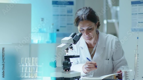 Mature female scientist in white coat using compound microscope and making notes, conducting chemistry research in laboratory