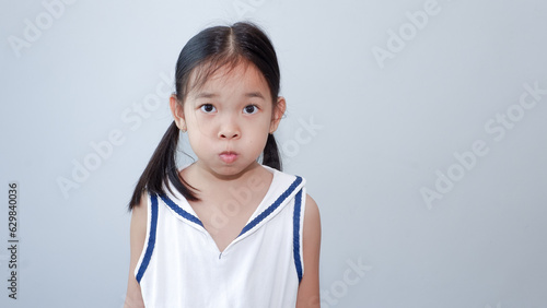 Portrait of small Asian female kid blows cheeks and puffs breath, has attractive look and pigtails hairstyle, keeps silence, wears casual white t shirt, isolated over light gray wall.