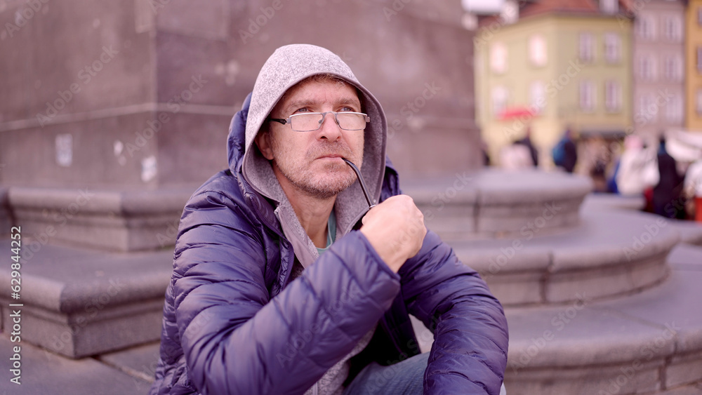Portrait of adult man with glasses sitting in the hood on square and smoking a tobacco pipe in the Palace Square, Warsaw Old Town Portrait of adult man with glasses sitting in the hood on square and smoking a tobacco pipe in the Palace Square, Warsaw Old Town