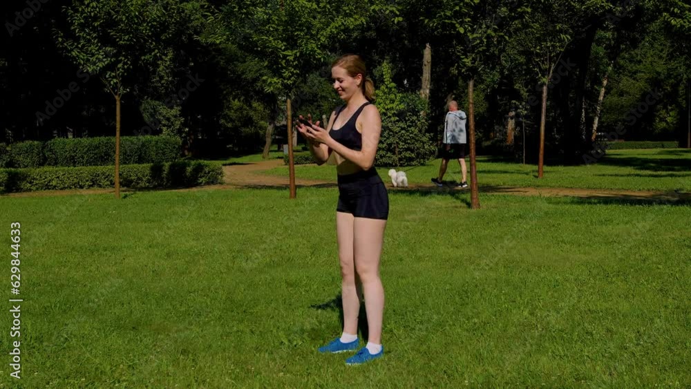 Positive multitasking attractive sporty fitness woman doing squat workout, networking and browsing social media content online on cellphone while practicing sports training on park lawn.