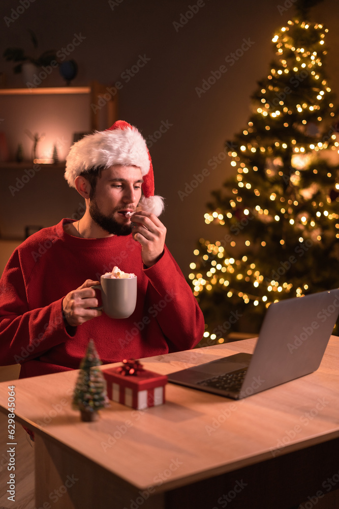 Young man at home office drinking cocoa and watching movie using laptop sitting at table in the evening on christmas eve