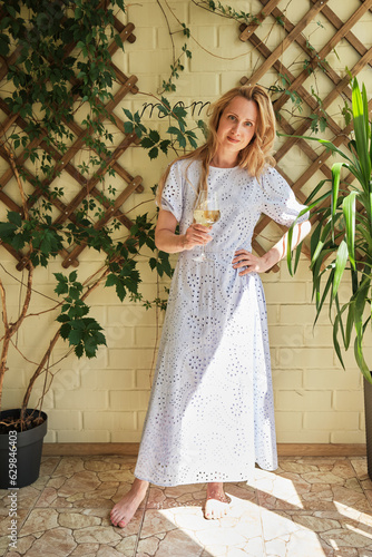 beautiful young white woman in a long blue light dress with long hair in a full-length standing on a terrace surrounded by green plants with a glass of white wine on a sunny summer day in a pensive po