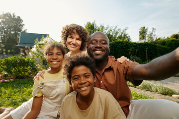Happy interracial family looking at camera with smiles while young African American man with smartphone taking selfie of his wife and their two kids