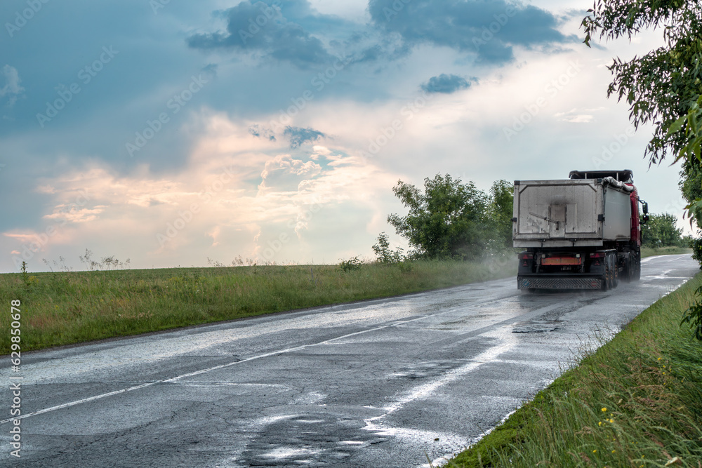 Heavy truck vehicle driving wet asphalt road driveway in stormy rainy weather with epic clouds and green landscape. Rear car view with water mist on wheels Heavy truck vehicle driving wet asphalt road driveway in stormy rainy weather with epic clouds and green landscape. Rear car view with water mist on wheels