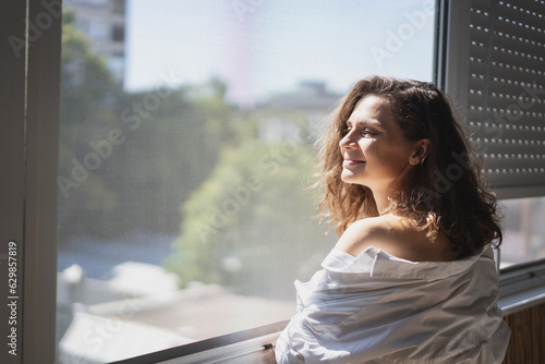 Young smiling caucasian woman in a white shirt standing on the balcony in the morning meeting a new day. View of summer city