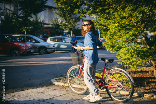 A young Caucasian woman using a phone while standing next to her bicycle in the summer city street.