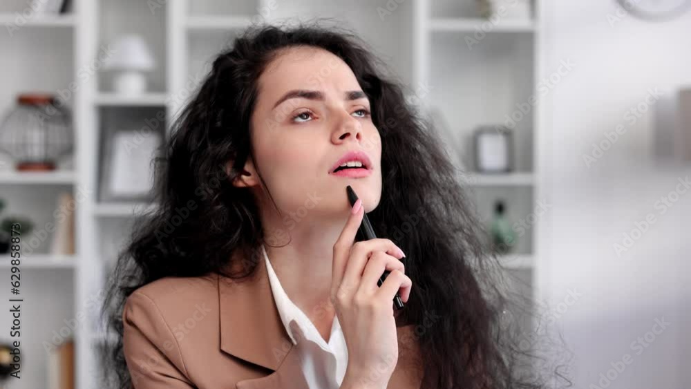 Young curly brunette woman, thoughtful looking for an idea, sits at a table and works on a laptop in a home bright office, looks at the camera. Programmer, remote work, work from home. Young curly brunette woman, thoughtful looking for an idea, sits at a table and works on a laptop in a home bright office, looks at the camera. Programmer, remote work, work from home.