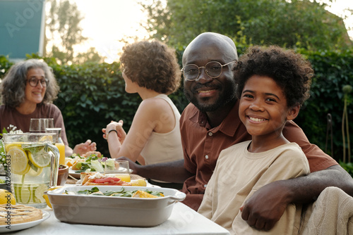 Happy young African American father and cute son sitting by table with homemade food and looking at camera against two women talking